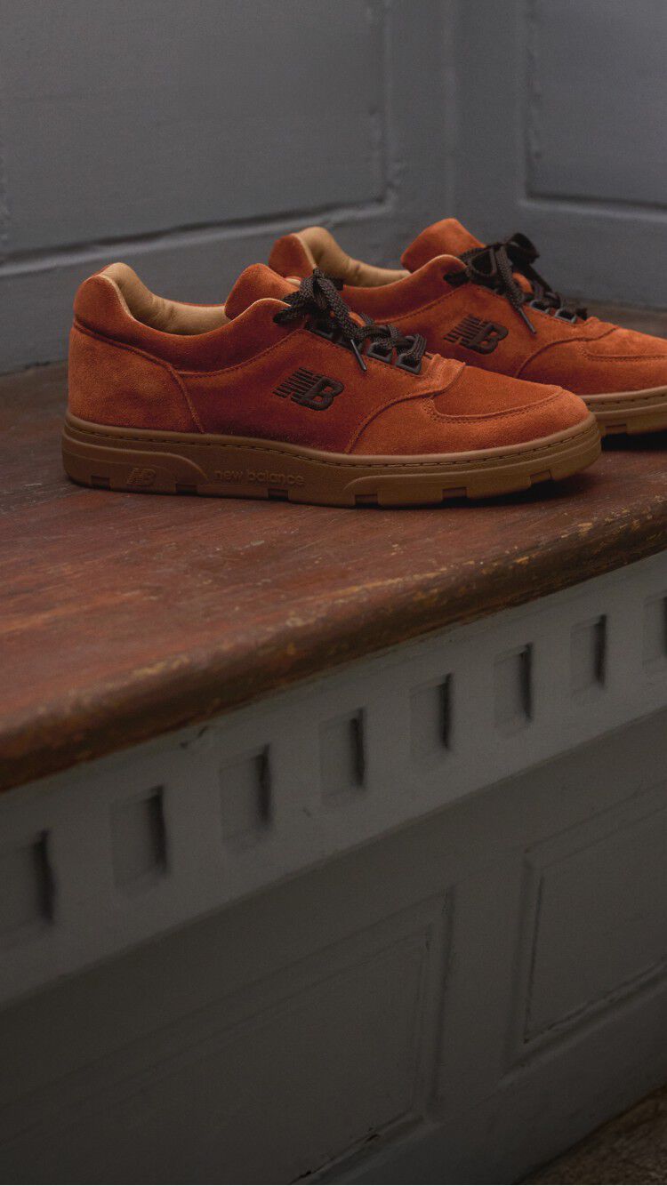 A pair of New Balance Made in UK sneakers sitting on a wooden window seat with panel detailing on the walls.   
